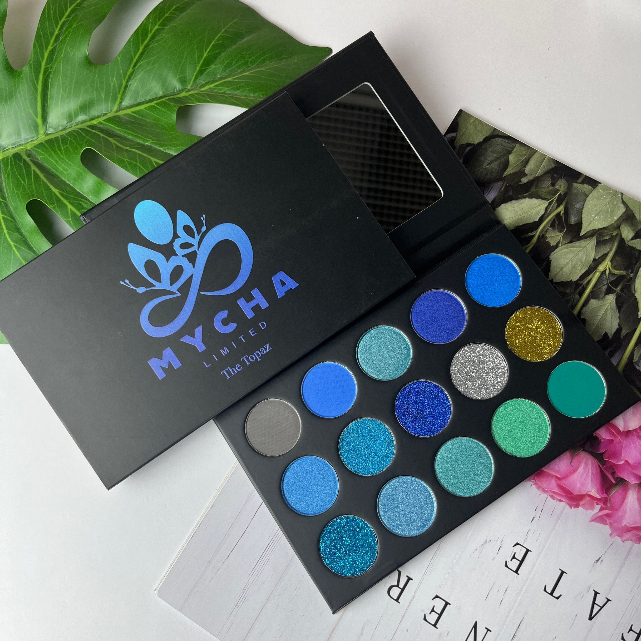 Open Topaz eyeshadow palette with various blue and glitter shades in black casing and MYCHA branding next to green leaf and floral decor