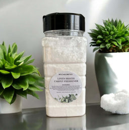 Linen Breeze Carpet Freshener bottle with white powder next to green potted plants on a gray surface