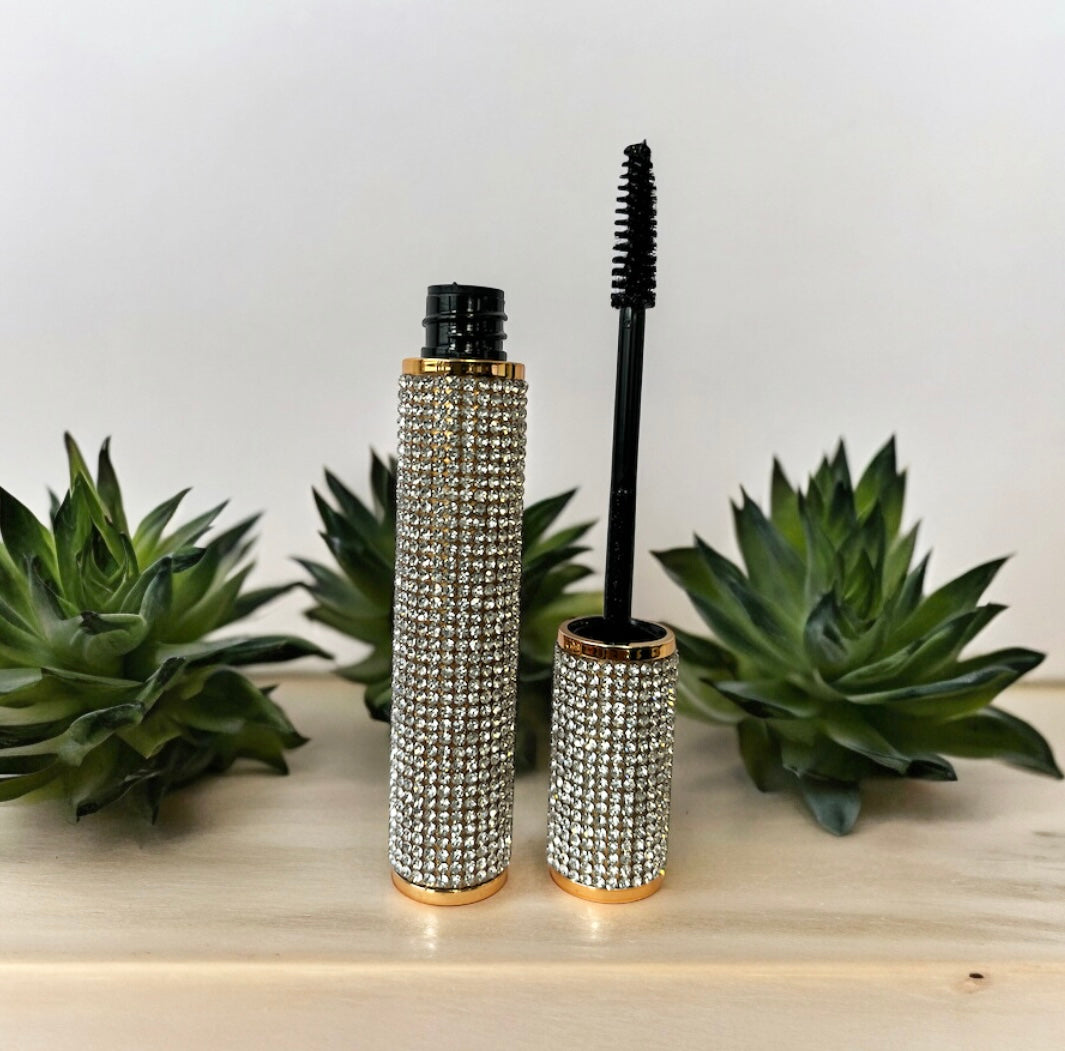 Waterproof 4D mascara tube and wand with rhinestone-studded gold casing on a wooden surface with green succulent plants in the background