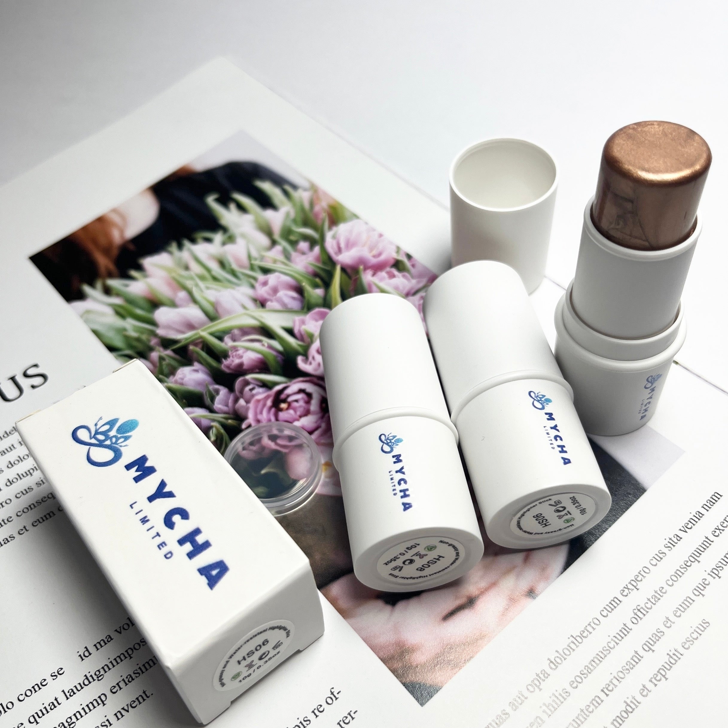 Radiant Aura Highlighter Stick in white tube with gold shimmer, displayed alongside packaging and floral magazine background.