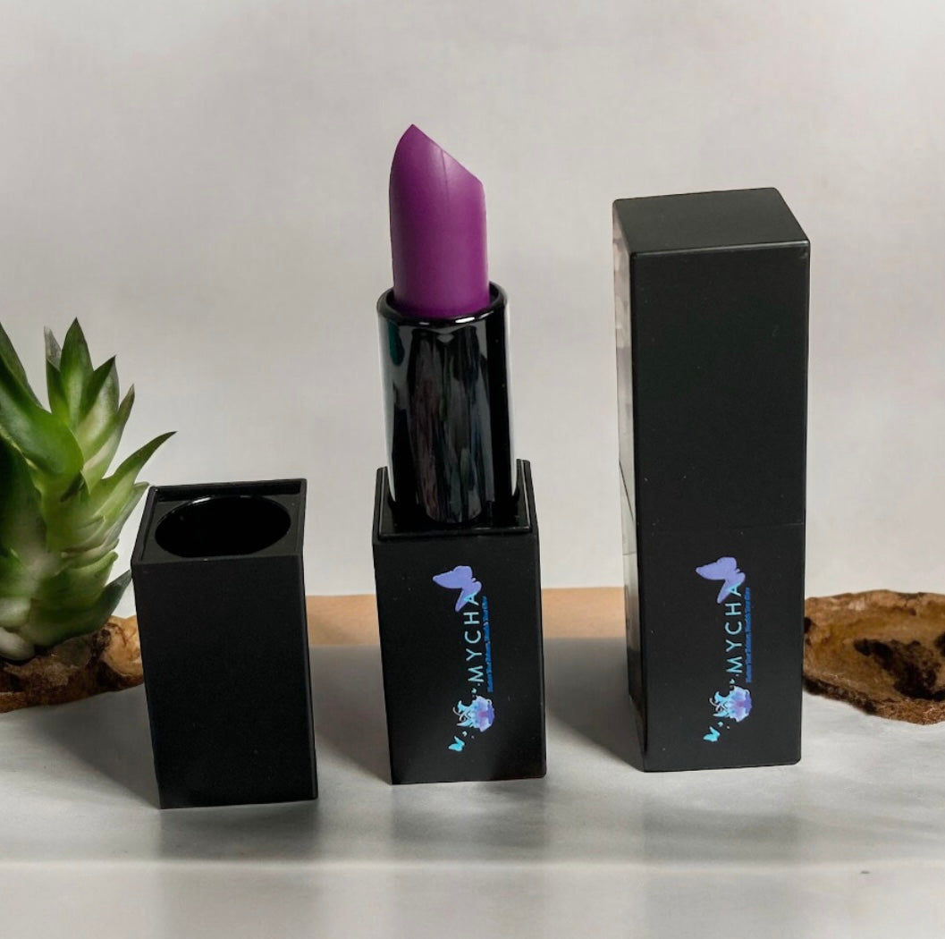 Silk Touch Matte Lipstick in vibrant purple shade with sleek black packaging and box on a white surface with green plant background