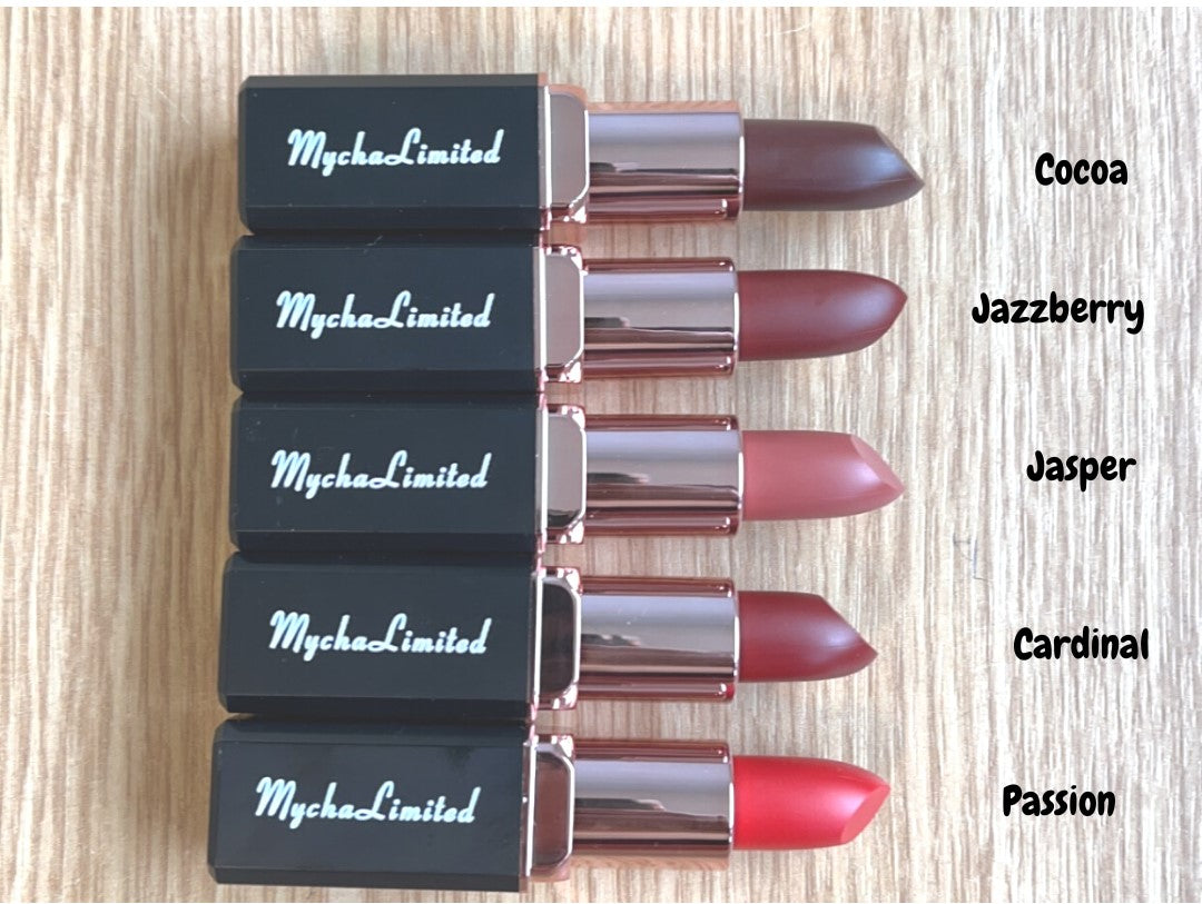 Set of five MychaLimited Enchanté Matte Lip Perfection lipsticks in shades Cocoa, Jazzberry, Jasper, Cardinal, and Passion with rose gold and black casing on wooden background